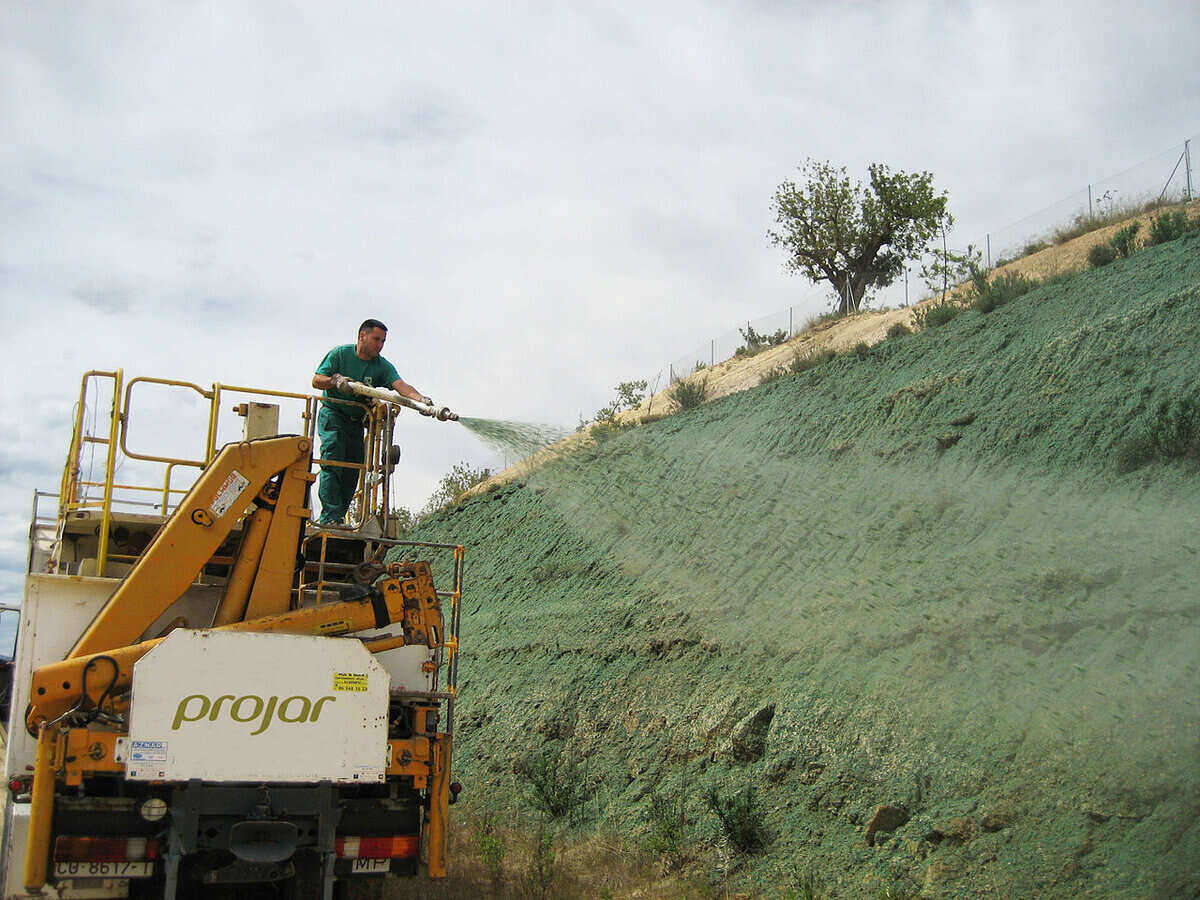 Man standing on top of a truck and uses a hose to spray for hydroseeding along the side of a steep incline